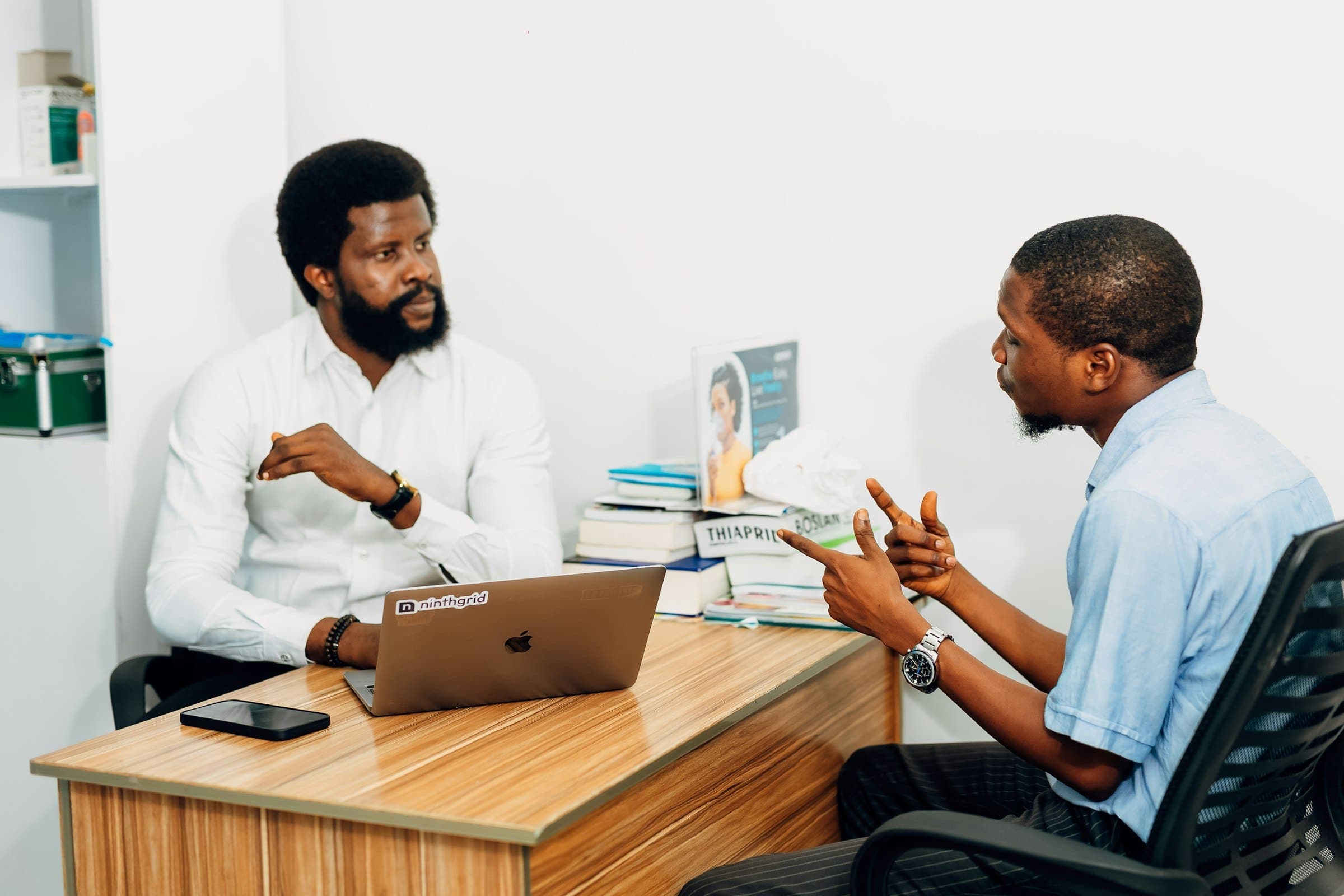 Two men in discussion across an office desk with a laptop and documents